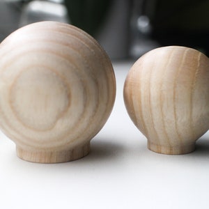 May include: Two wooden spheres, one larger than the other, with a natural wood grain finish. The spheres are smooth and round, and they are both sitting on a white surface.