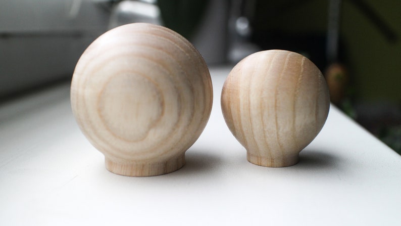 May include: Two wooden spheres, one larger than the other, with a natural wood grain finish. The spheres are smooth and round, and they are both sitting on a white surface.