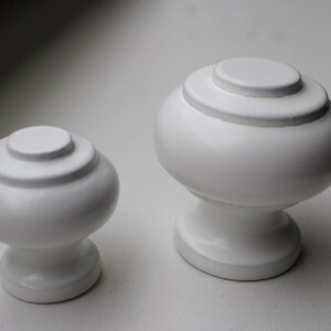 May include: Two white ceramic cabinet knobs with a rounded, slightly bulbous shape. The larger knob has a diameter of approximately 5 cm, while the smaller knob is about 4 cm in diameter. Both knobs have a smooth, glossy finish.