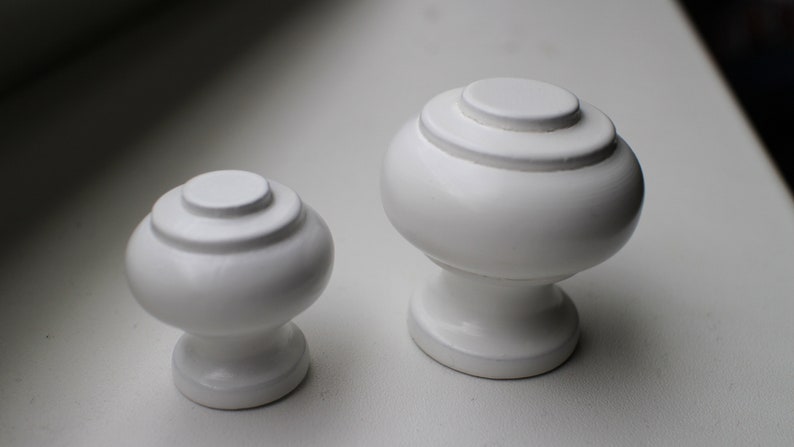 May include: Two white ceramic cabinet knobs with a rounded, slightly bulbous shape. The larger knob has a diameter of approximately 5 cm, while the smaller knob is about 4 cm in diameter. Both knobs have a smooth, glossy finish.