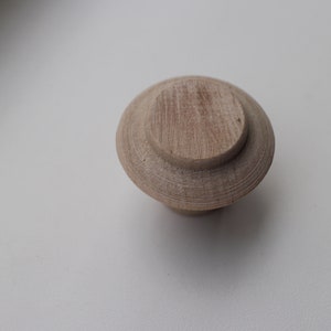 Op de afbeelding: Een houten knop met een ronde bovenkant en een licht verhoogde rand. De knop is lichtbruin van kleur en heeft een gladde afwerking.