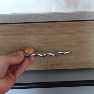 Pu&ograve; includere: Una maniglia per armadio con una maniglia ondulata color argento e un piccolo pomello rotondo marrone chiaro. La maniglia &egrave; montata su un'anta di un armadio in legno marrone chiaro.