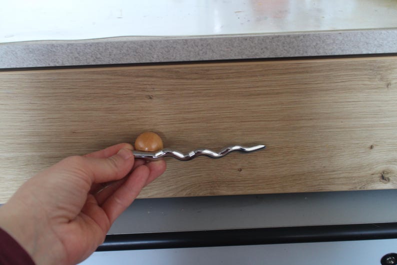 Pu&ograve; includere: Una maniglia per armadio con una maniglia ondulata color argento e un piccolo pomello rotondo marrone chiaro. La maniglia &egrave; montata su un'anta di un armadio in legno marrone chiaro.
