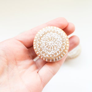 May include: A hand holding a round wooden knob with a white mandala design painted on it.