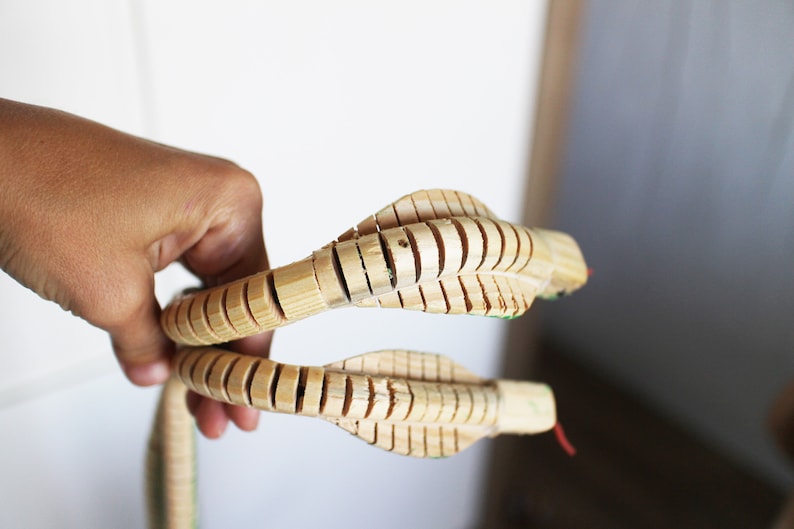 Puede incluir: Un juguete de serpiente de madera con partes del cuerpo segmentadas, hecho de madera natural. La serpiente est&aacute; enroscada y tiene dos cabezas.
