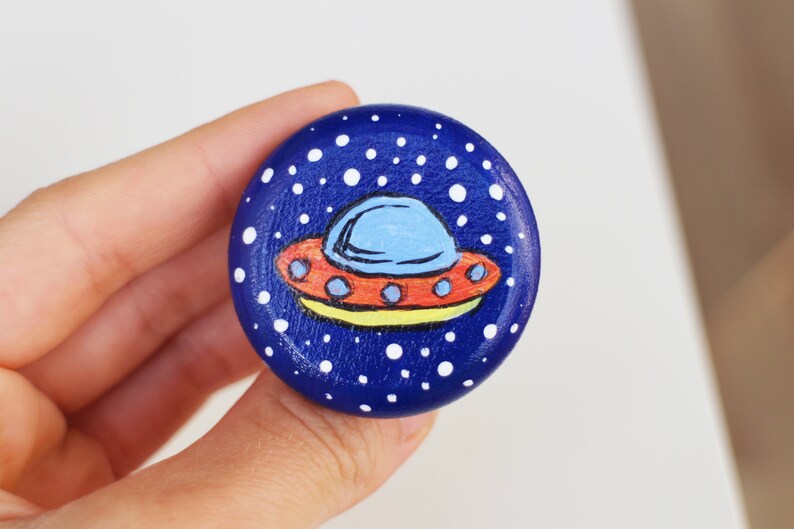 May include: A hand-painted wooden button with a blue background and white dots, depicting a spaceship. The spaceship is orange, blue, and yellow. The button is round.