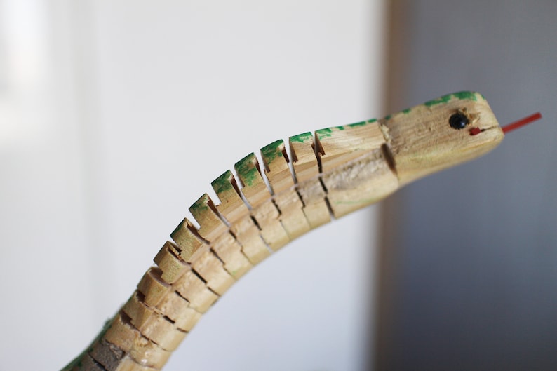 Puede incluir: Un juguete de serpiente de madera con escamas pintadas de verde y una lengua roja. La serpiente est&aacute; hecha de piezas de madera individuales que est&aacute;n conectadas entre s&iacute;.