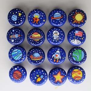 A collection of sixteen round, blue wooden knobs, each hand-painted with a different space-themed design. The designs include planets, rockets, astronauts, stars, and the American flag on the moon. White dots represent stars.
