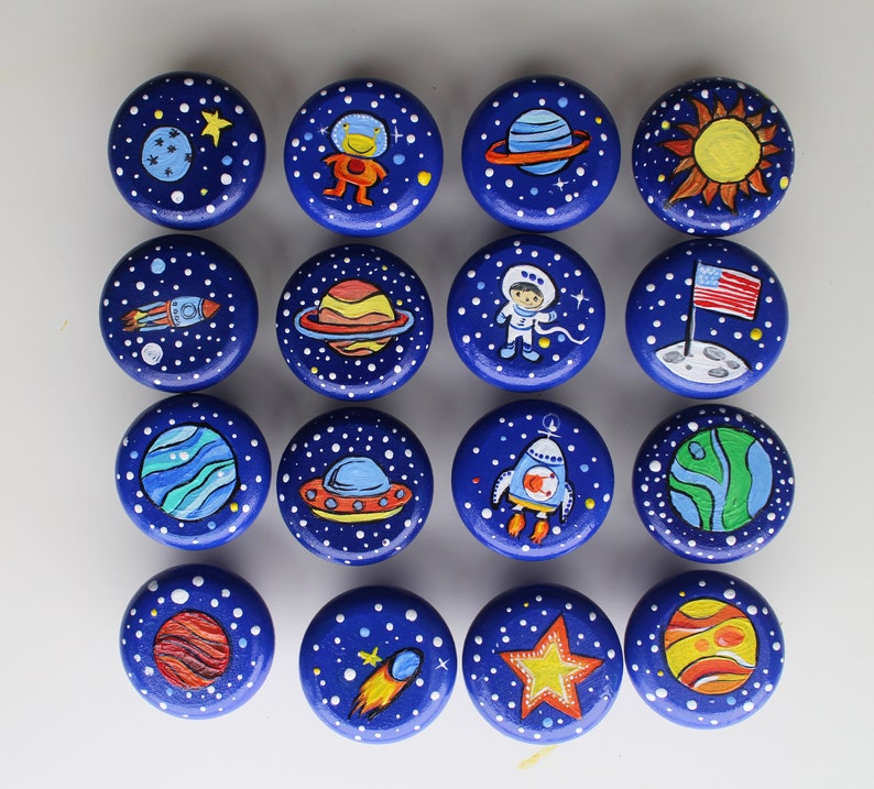 A collection of sixteen round, blue wooden knobs, each hand-painted with a different space-themed design. The designs include planets, rockets, astronauts, stars, and the American flag on the moon. White dots represent stars.