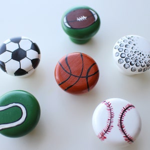 May include: Six round wooden knobs painted to look like different sports balls: a soccer ball, a football, a basketball, a golf ball, a tennis ball, and a baseball.