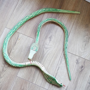 Puede incluir: Una serpiente de juguete de madera verde y blanca con un cuerpo segmentado y una lengua roja. La serpiente est&aacute; enroscada en un suelo de madera.