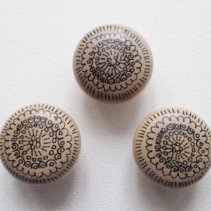 Peut inclure: Trois boutons ronds en bois avec des motifs de mandalas noirs. Les boutons sont peints d'une couleur brun clair et ont une finition lisse et polie.