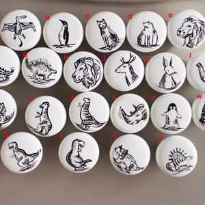 May include: Set of 18 white ceramic knobs with black ink illustrations of various animals, including a penguin, a lion, a deer, a rabbit, an elephant, a zebra, a rhinoceros, and several dinosaurs.