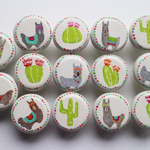 Può includere: Dodici pomelli rotondi in legno con disegni dipinti. I disegni includono lama, cactus e motivi geometrici in colori vivaci come rosa, verde, blu e giallo.
