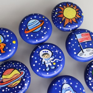 May include: A collection of hand-painted wooden knobs with a space theme. Each knob is a deep blue with white star details and features planets, astronauts, and a sun. The knobs are round and have a diameter of approximately 1.5 inches.