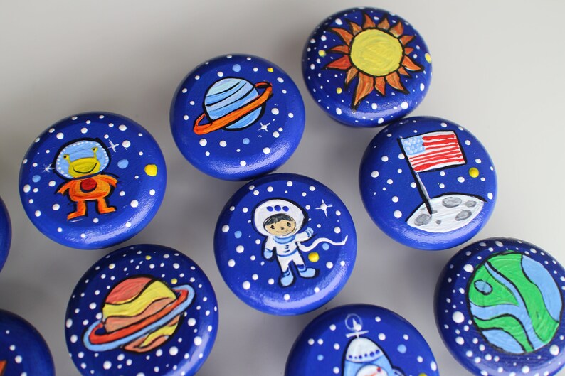 May include: A collection of hand-painted wooden knobs with a space theme. Each knob is a deep blue with white star details and features planets, astronauts, and a sun. The knobs are round and have a diameter of approximately 1.5 inches.