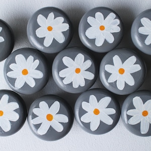 Puede incluir: Doce pomos redondos grises con diseños de margaritas blancas y centros amarillos.