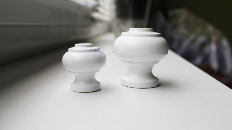 May include: Two white ceramic finials, one larger than the other, with a rounded top and a slightly flared base.