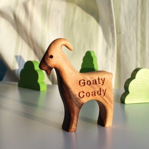May include: A wooden goat figurine with the text "Goaty Coady" carved on its side. The goat is brown with a curved horn and stands on a light blue surface. Green wooden tree and bush cutouts are in the background.