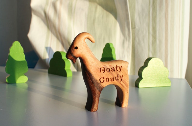 May include: A wooden goat figurine with the text "Goaty Coady" carved on its side. The goat is brown with a curved horn and stands on a light blue surface. Green wooden tree and bush cutouts are in the background.