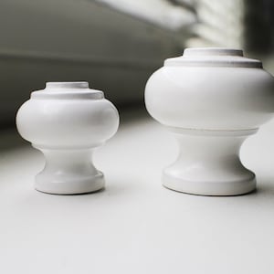 Könnte beinhalten: Zwei weiße Keramik-Abschlusskappen, eine größer als die andere, mit abgerundeter Oberseite und einer Basis. Die Abschlusskappen befinden sich auf einer weißen Oberfläche.