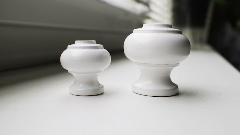 May include: Two white ceramic finials, one larger than the other, with a rounded top and a base. The finials are on a white surface.