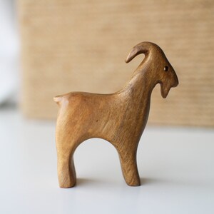 May include: A handcrafted wooden goat figurine with a light brown finish. The goat has curved horns, a small tail, and is standing on four legs. The figurine is likely a decorative item or a toy.