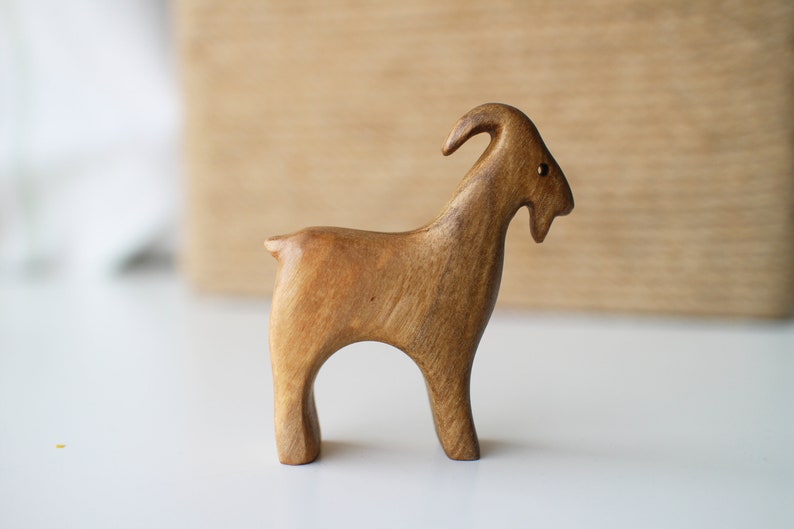 May include: A handcrafted wooden goat figurine with a light brown finish. The goat has curved horns, a small tail, and is standing on four legs. The figurine is likely a decorative item or a toy.