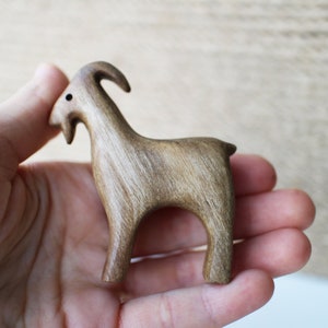 May include: A small, wooden goat figurine with a curved horn and a short tail. The goat is carved from light brown wood and has a smooth finish. The figurine is held in a person's hand.