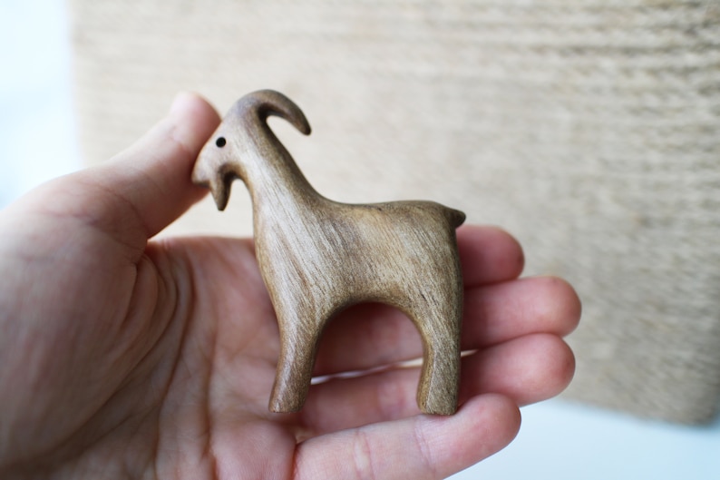 May include: A small, wooden goat figurine with a curved horn and a short tail. The goat is carved from light brown wood and has a smooth finish. The figurine is held in a person's hand.