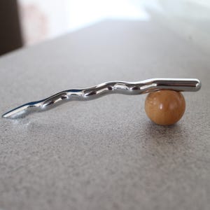 Pu&ograve; includere: Un oggetto metallico argentato e ondulato in equilibrio su una piccola sfera di legno rotonda. Il pezzo di metallo ha una superficie liscia e riflettente e un'estremit&agrave; appuntita. La sfera &egrave; di colore marrone chiaro.
