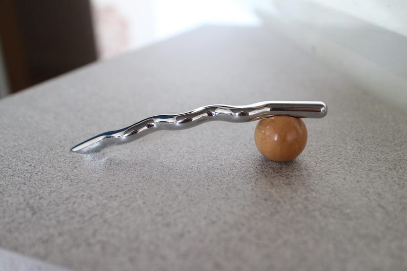 Pu&ograve; includere: Un oggetto metallico argentato e ondulato in equilibrio su una piccola sfera di legno rotonda. Il pezzo di metallo ha una superficie liscia e riflettente e un'estremit&agrave; appuntita. La sfera &egrave; di colore marrone chiaro.
