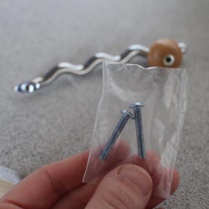 Pu&ograve; includere: Una maniglia per mobili con una maniglia ondulata color argento e un'estremit&agrave; arrotondata marrone chiaro. Due viti argentate sono in un piccolo sacchetto di plastica trasparente. L'hardware &egrave; probabilmente per un cassetto o un armadio.