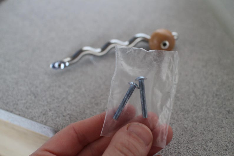 Pu&ograve; includere: Una maniglia per mobili con una maniglia ondulata color argento e un'estremit&agrave; arrotondata marrone chiaro. Due viti argentate sono in un piccolo sacchetto di plastica trasparente. L'hardware &egrave; probabilmente per un cassetto o un armadio.