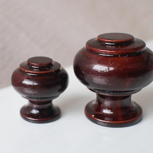 Op de afbeelding: Twee donkerbruine houten kastknoppen met een glanzende afwerking. De knoppen hebben een afgerond, gelaagd ontwerp en staan op een wit oppervlak. De ene knop is kleiner dan de andere.