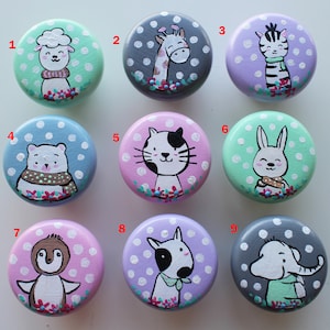 Może przedstawiać: Dziewięć drewnianych gałek z malowanymi wzorami zwierząt. Każda gałka przedstawia inne zwierzę, w tym jagnię, żyrafę, zebrę, niedźwiedzia polarnego, kota, królika, pingwina, bulteriera i słonia. Gałki są pomalowane w różnych kolorach, w tym zielonym, szarym, fioletowym, niebieskim, różowym i białym. Każda gałka ma białe kropki na tle.