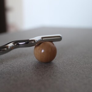Pu&ograve; includere: Una barra di metallo argentato lucido poggia su una piccola sfera di legno rotonda. La barra ha un design curvo e un'estremit&agrave; piatta. La sfera &egrave; marrone chiaro. Gli oggetti sono su una superficie grigia.