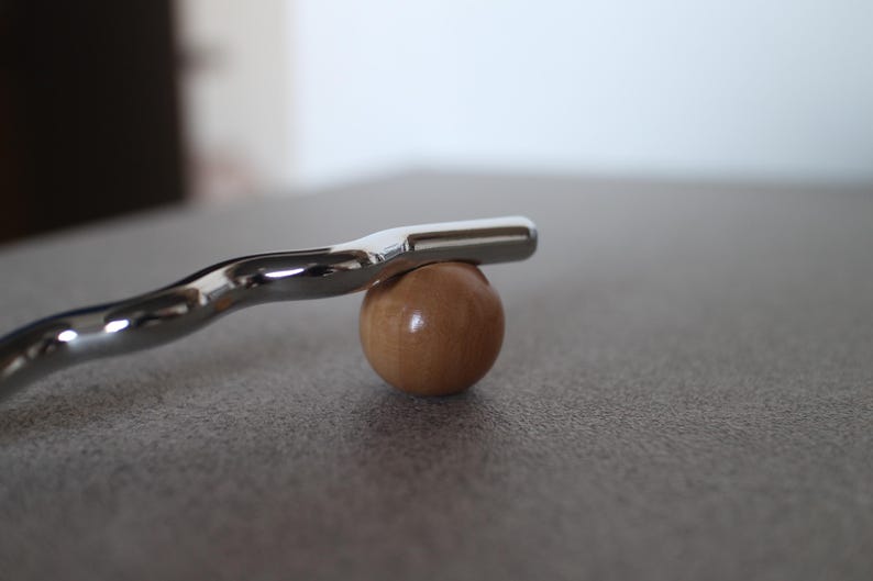 Pu&ograve; includere: Una barra di metallo argentato lucido poggia su una piccola sfera di legno rotonda. La barra ha un design curvo e un'estremit&agrave; piatta. La sfera &egrave; marrone chiaro. Gli oggetti sono su una superficie grigia.