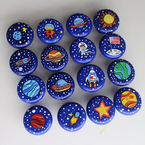 A collection of hand-painted, round cabinet knobs with a space theme. Each knob features a dark blue background with white stars and various space-related illustrations, including planets, rockets, and astronauts. The knobs are designed for furniture.