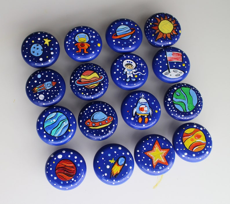 A collection of hand-painted, round cabinet knobs with a space theme. Each knob features a dark blue background with white stars and various space-related illustrations, including planets, rockets, and astronauts. The knobs are designed for furniture.