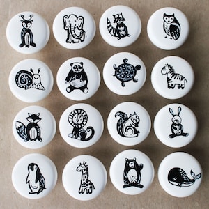 May include: Set of 15 white ceramic knobs with black animal illustrations. The knobs feature a variety of animals, including a deer, elephant, raccoon, owl, snail, panda, turtle, zebra, fox, lion, squirrel, rabbit, penguin, giraffe, and bear.