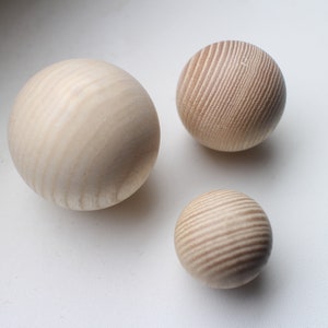 May include: Three wooden spheres of varying sizes, all with a light brown color and a wood grain pattern. The spheres are arranged on a white surface.
