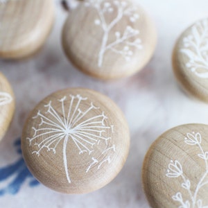 Può includere: Sei pomelli rotondi in legno con disegni floreali bianchi dipinti su di essi. I pomelli sono disposti a semicerchio su una superficie bianca.