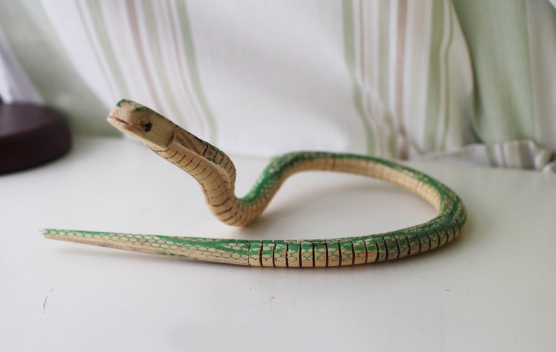 Puede incluir: Un juguete de serpiente de madera pintado en verde y marr&oacute;n. La serpiente est&aacute; enroscada en un c&iacute;rculo con la cabeza levantada.