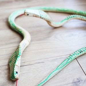 Puede incluir: Una serpiente de juguete de madera con escamas pintadas de verde y blanco. La serpiente est&aacute; enroscada en forma de c&iacute;rculo con la cabeza mirando hacia la c&aacute;mara.