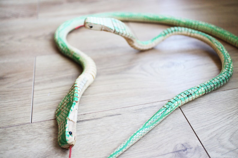 Puede incluir: Una serpiente de juguete de madera con escamas pintadas de verde y blanco. La serpiente est&aacute; enroscada en forma de c&iacute;rculo con la cabeza mirando hacia la c&aacute;mara.