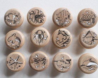 May include: Twelve wooden knobs with black and white illustrations of sea creatures, including a jellyfish, a crab, a starfish, a turtle, a fish, an octopus, a narwhal, a dolphin, and a shark.