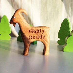 May include: A wooden goat figurine with the text "Goaty Coady" carved into its side. The goat is brown and stands on four legs. Green wooden tree and bush cutouts are in the background.