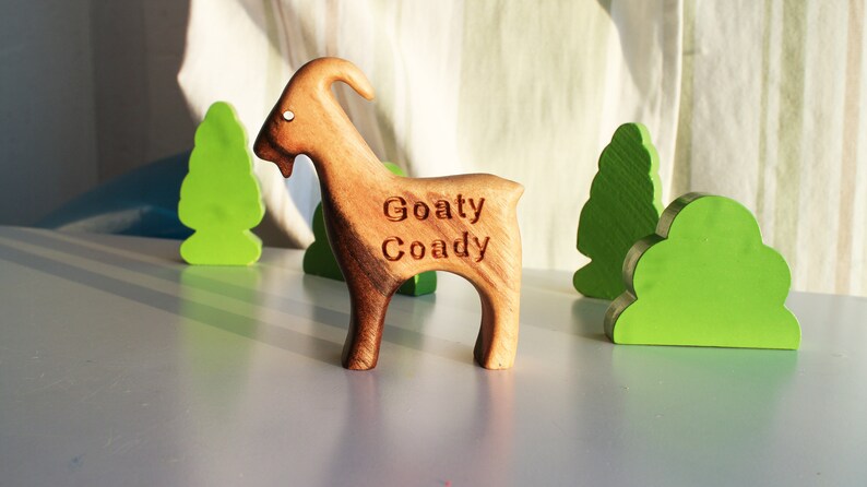 May include: A wooden goat figurine with the text "Goaty Coady" carved into its side. The goat is brown and stands on four legs. Green wooden tree and bush cutouts are in the background.