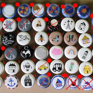 May include: A selection of hand-painted bottle stoppers with diverse designs. The stoppers showcase images of animals, buildings, and other decorative elements. The stoppers are round and painted in various colours, including blue, white, and black.